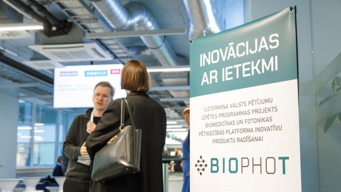 BioPhot plakāts ar uzrakstu "Inovācijas ar ietekmi" un divi cilvēki sarunājas