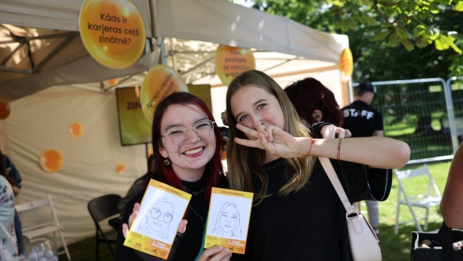 Jaunieši Liepājā festivālā KOPUMS pozē fotogrāfam ar karikatūrām, ko uzzīmējis MI robots