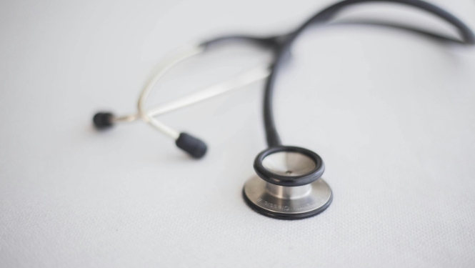 Stetoskops novietots uz gaišas virsmas. Attēls simbolizē veselības aprūpi, medicīnas pētniecību un pacientu aprūpi.