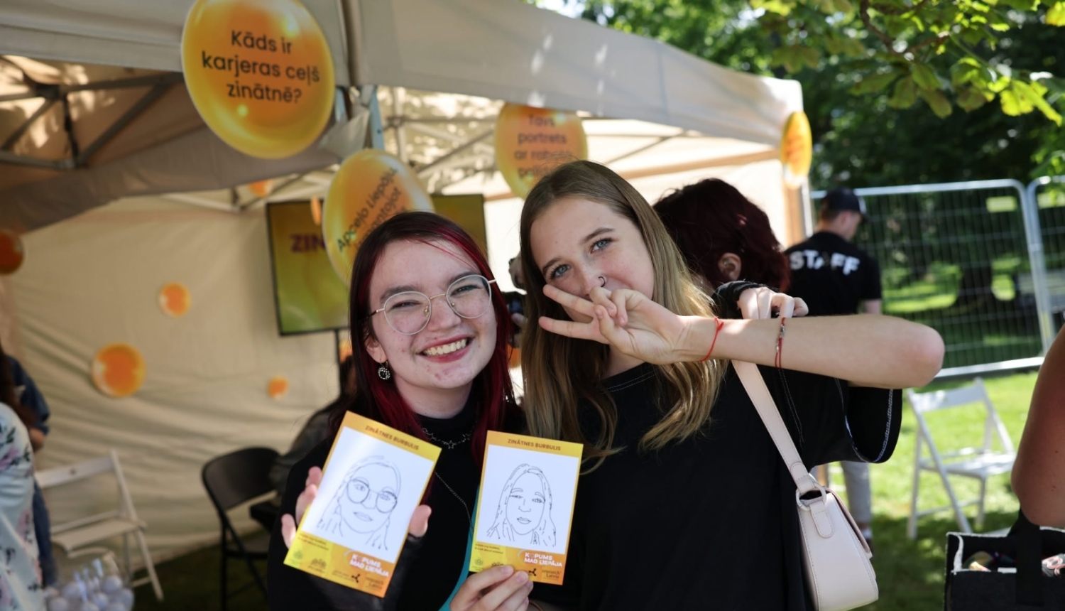 Jaunieši Liepājā festivālā KOPUMS pozē fotogrāfam ar karikatūrām, ko uzzīmējis MI robots
