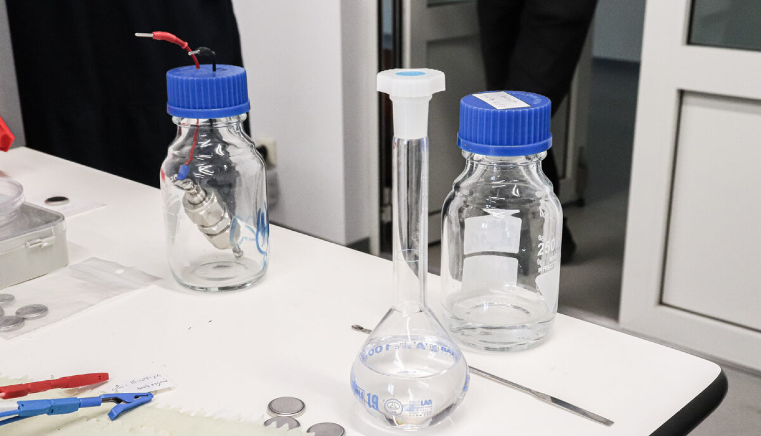 Laboratorijas galds, tukša kolba un pudelītes ar ziliem korķiem. 