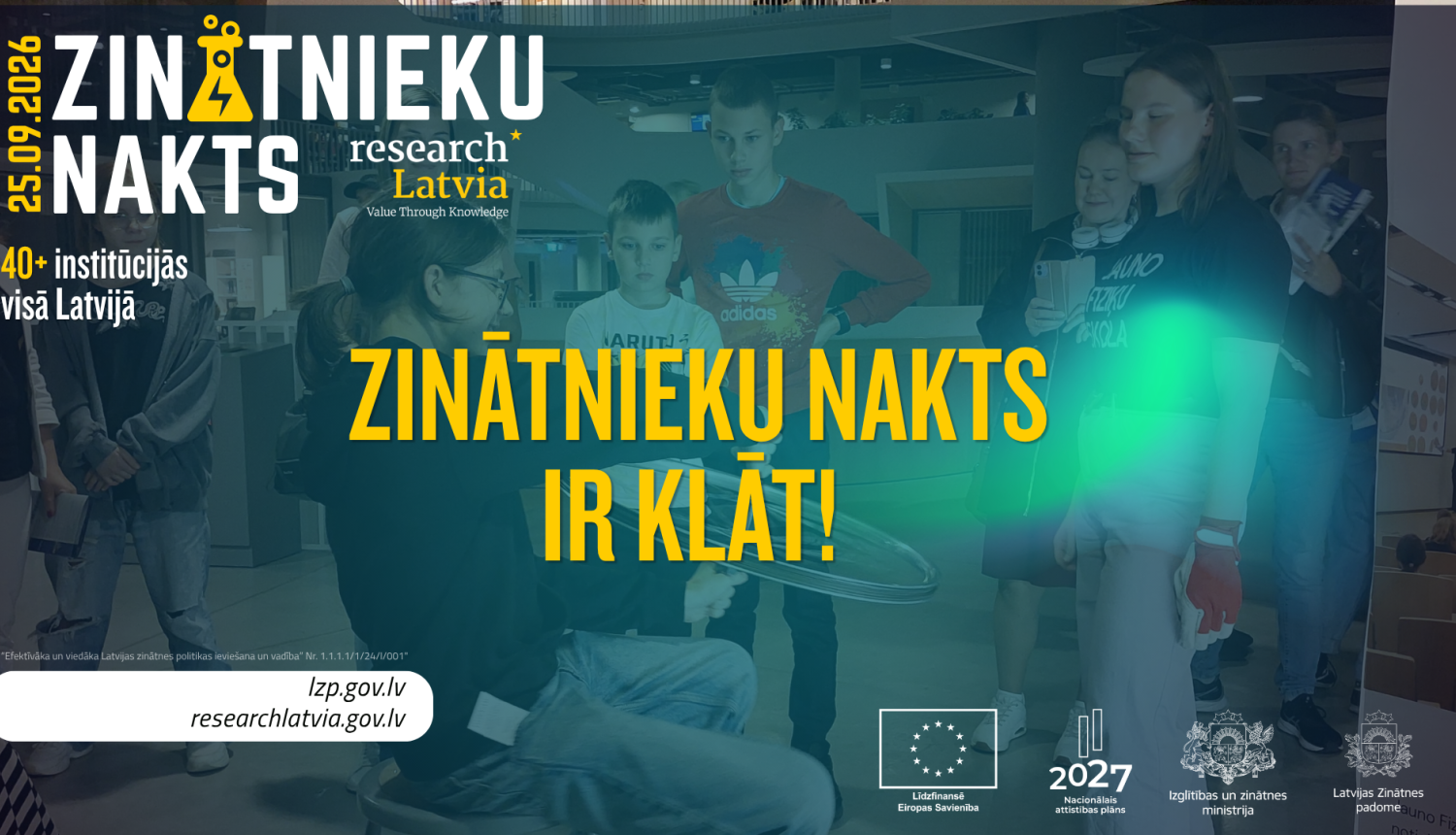 Zinātnieku nakts attēls ar logo un uzrakstu "Zinātnieku nakts ir klāt". Fonā bērni universitātes telpā piedalās aktivitātēs. 