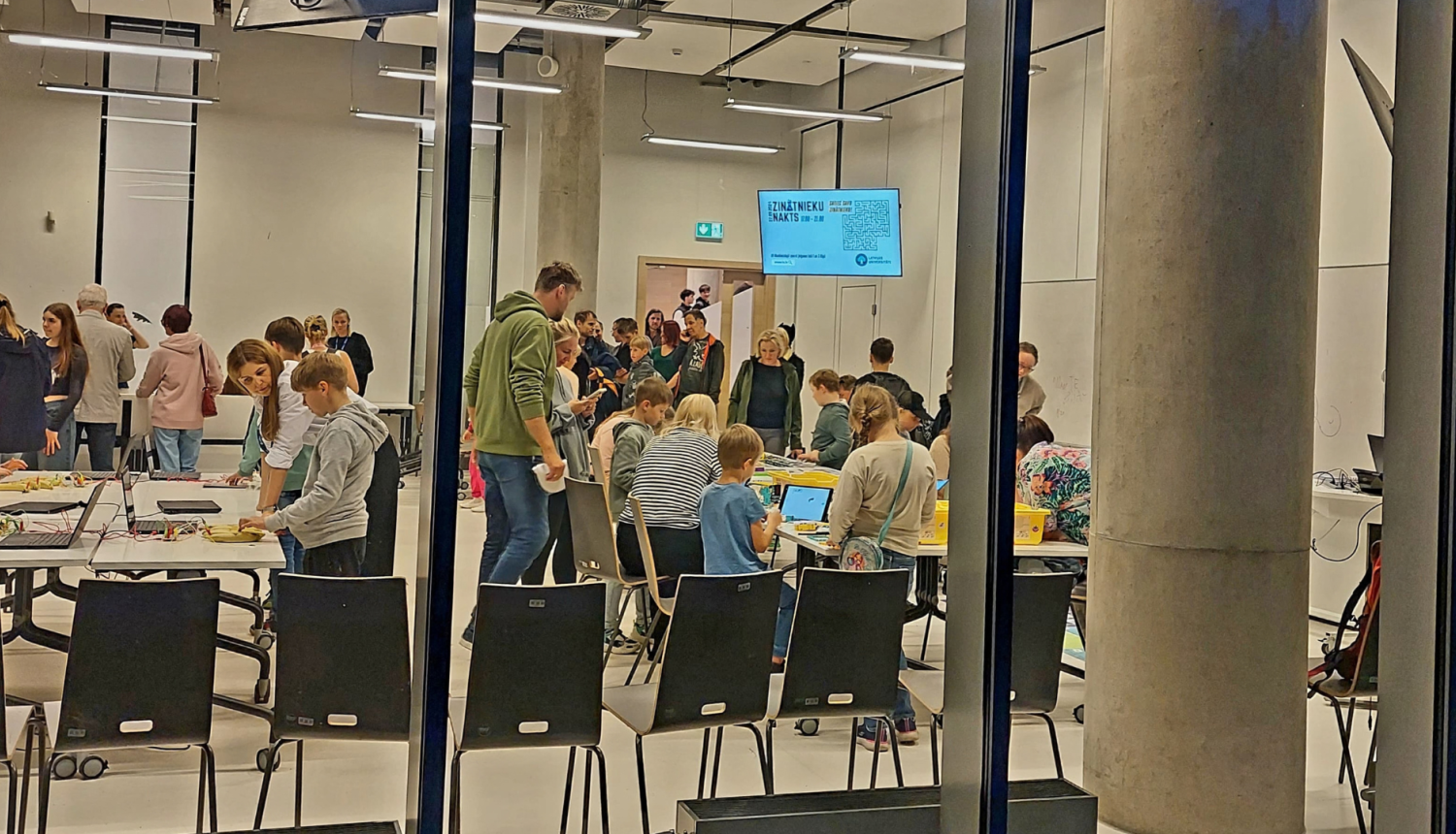 Caur logu redzami Zinātnieku nakts dalībnieki universitātes telpās. 