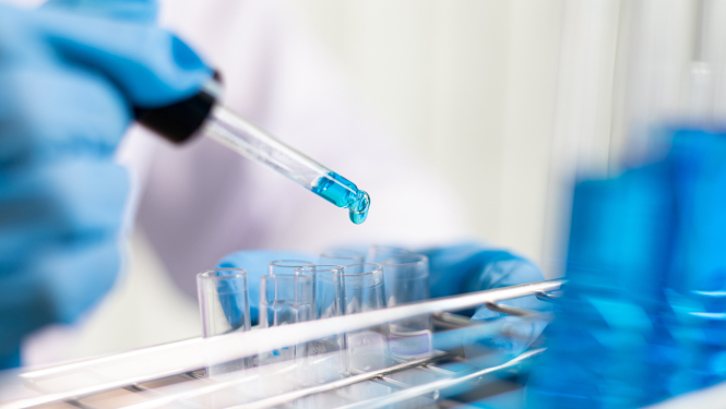 Laboratorijas pētnieks aizsargcimdos ar pilinātāju iepilina zilu šķidrumu stikla mēģenē – zinātniskais darbs laboratorijas apstākļos.