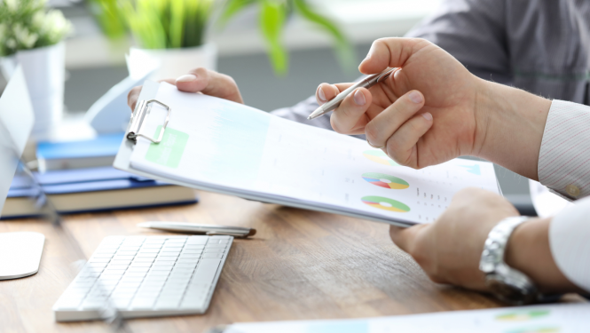 Cilvēks, sēžot pie galda, tur rokās mapi un pildspalvu