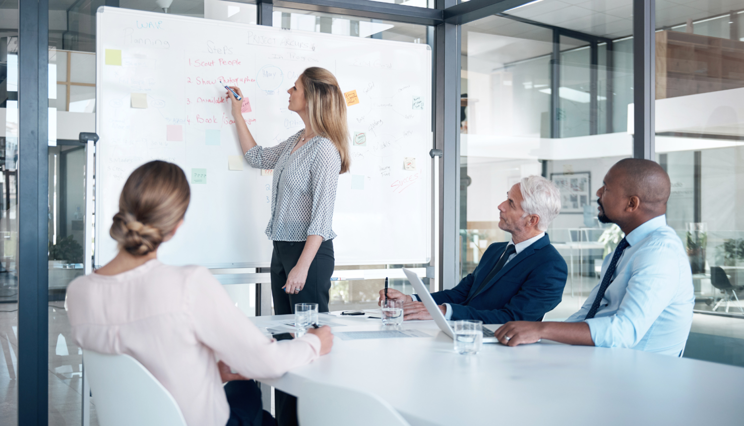 Projektu vadītājs sapulču telpā prezentē projektu. Sapulces dalībnieki sēž pie galda