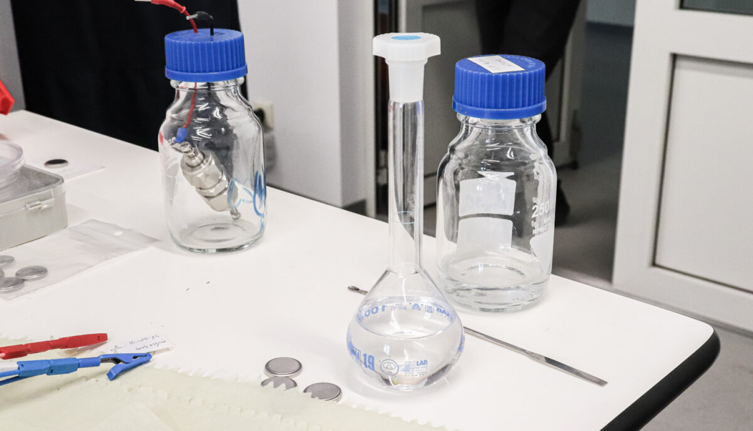 Laboratorijas galds, tukša kolba un pudelītes ar ziliem korķiem. 