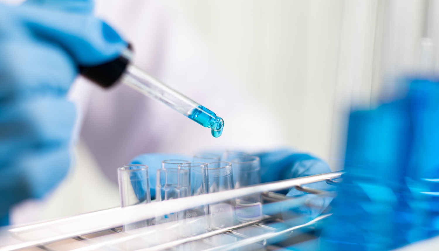 Laboratorijas pētnieks aizsargcimdos ar pilinātāju iepilina zilu šķidrumu stikla mēģenē – zinātniskais darbs laboratorijas apstākļos.